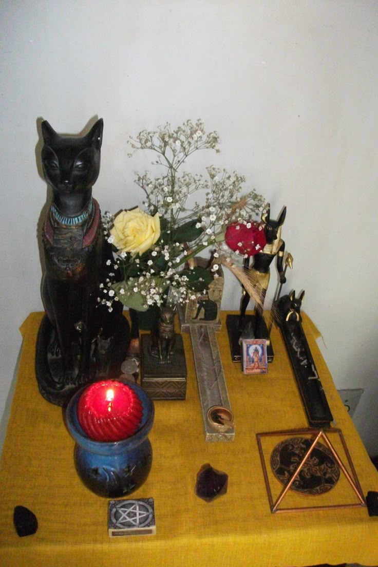 a yellow table topped with two black cats and other items next to a vase filled with flowers