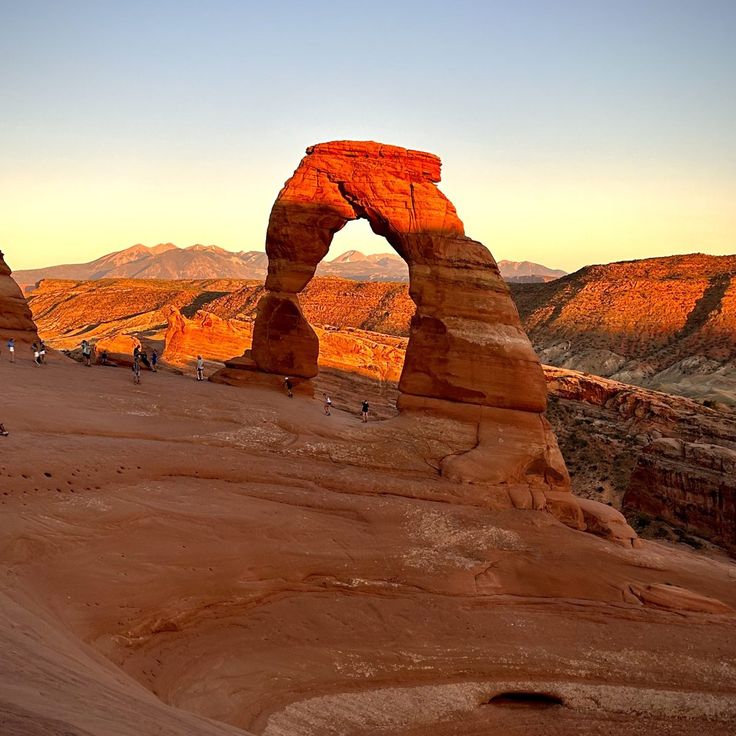 an arch shaped rock formation in the desert with people walking around it and mountains in the background