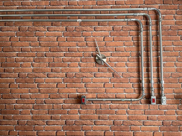 a brick wall with metal pipes and a clock on it's side, in front of a red brick wall