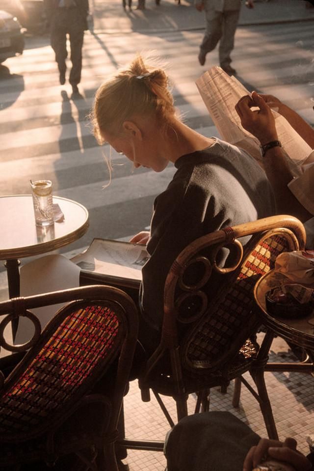 a woman is sitting at an outdoor table with her cell phone in her hand and people walking by