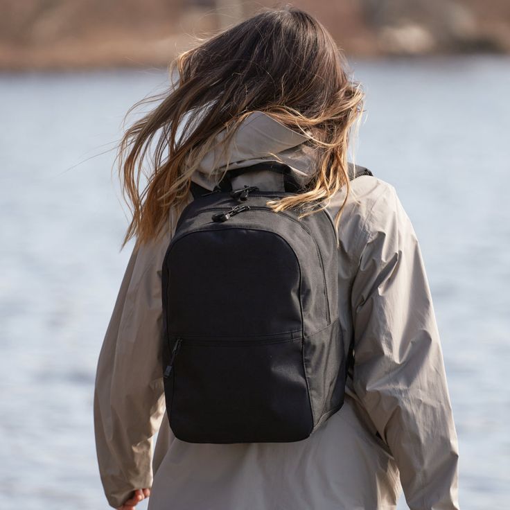 a woman with her back to the camera, walking by water wearing a black backpack