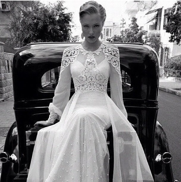 a woman sitting on the back of a car wearing a long white dress with sheer sleeves