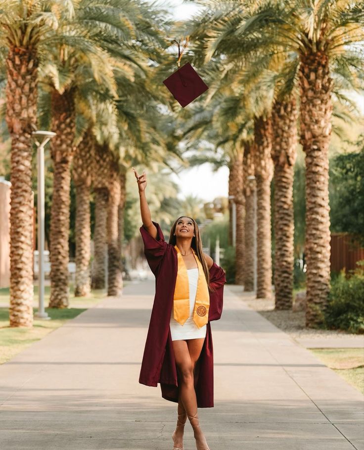 a woman wearing a graduation cap and gown is throwing her mortar in the air while standing on a path lined with palm trees