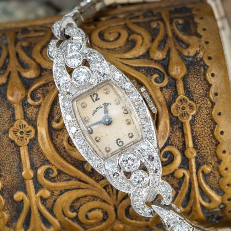 This beautiful circa 1950s Hamilton wristwatch features a platinum case, platinum lugs and a 14K white gold bracelet. The case and lugs are accented with four (4), bezel set, round brilliant cut diamonds and forty (40), bead set, round single cut diamonds. The 14K white gold bracelet is accented with twenty-six (26), bead set, round brilliant cut diamonds. The fold-over style clasp is accented with four (4), bead set, round single cut diamonds. The case measures 14mm wide (Case No. 5005217). The movement is a Hamilton manual wind, 17 jewel movement (Caliber 750). Serial No. 670874A. This wrist watch fits up to a 5.75 inch wrist. These beautiful old watches are typically for fashion rather than accurate time keeping. EraGem is not able to offer any warranty on watch movements. Luxury Engraved Vintage Diamond Watch, Luxury Classic Jewelry And Watches With Manual Winding, Luxury Antique Formal Watch Accessories, Luxury Vintage Watch Accessories With Stainless Steel Clasp, Luxury Vintage Watches With Quartz Movement, Vintage Diamond Watch With Diamond Hour Markers For Evening, Art Deco Platinum Watch For Anniversary, Timeless Platinum Diamond Watch For Wedding, Art Deco Platinum Anniversary Watch