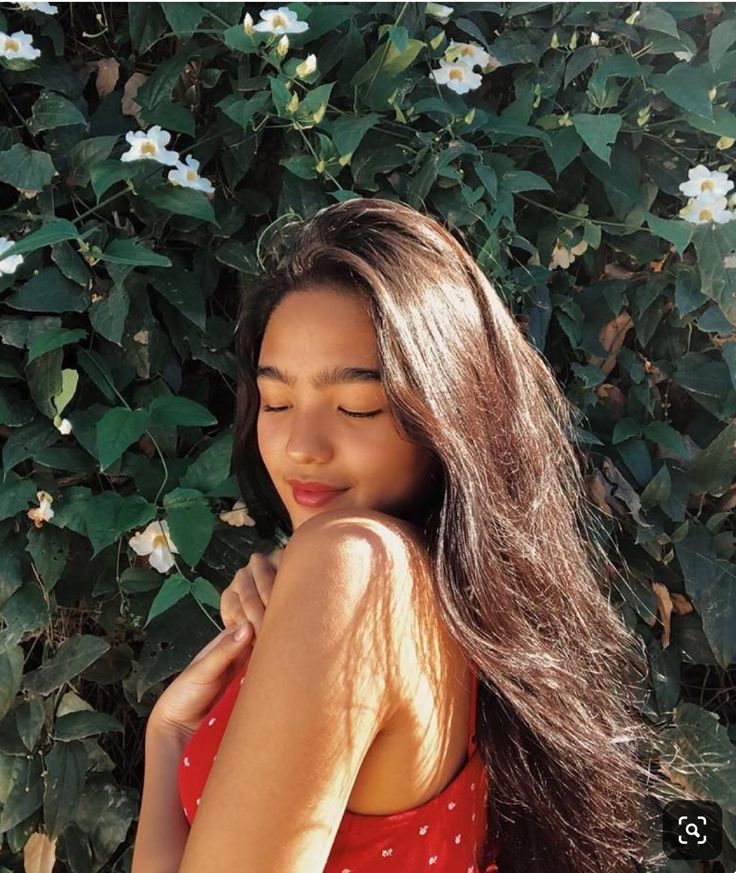 a woman with long hair standing in front of some bushes and flowers smiling at the camera