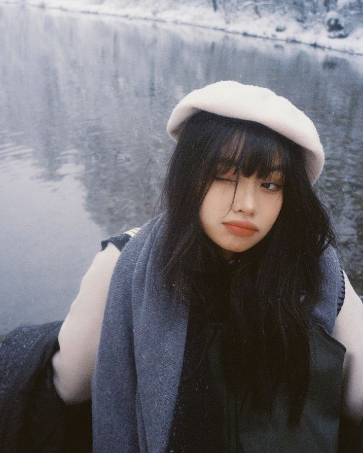 a woman with long black hair wearing a white hat and scarf over her shoulders standing in front of a body of water