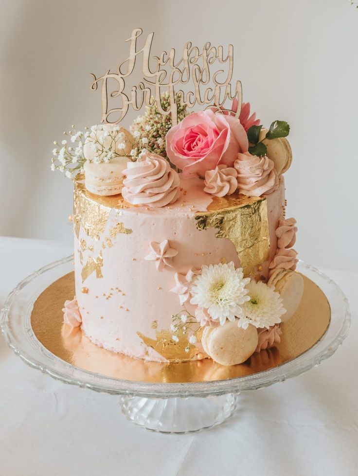 a pink and gold birthday cake with flowers on top is sitting on a glass platter