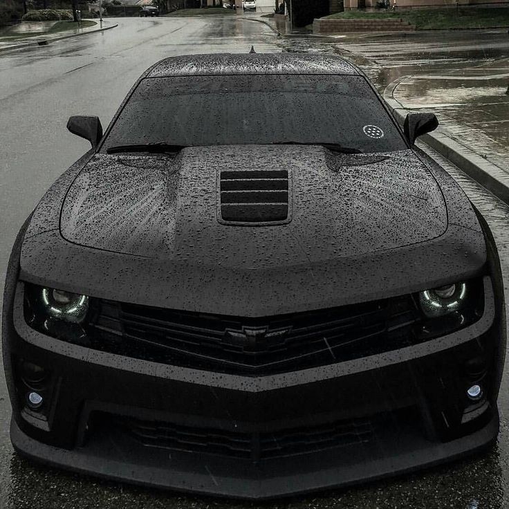 the front end of a car covered in water on a wet street with buildings behind it