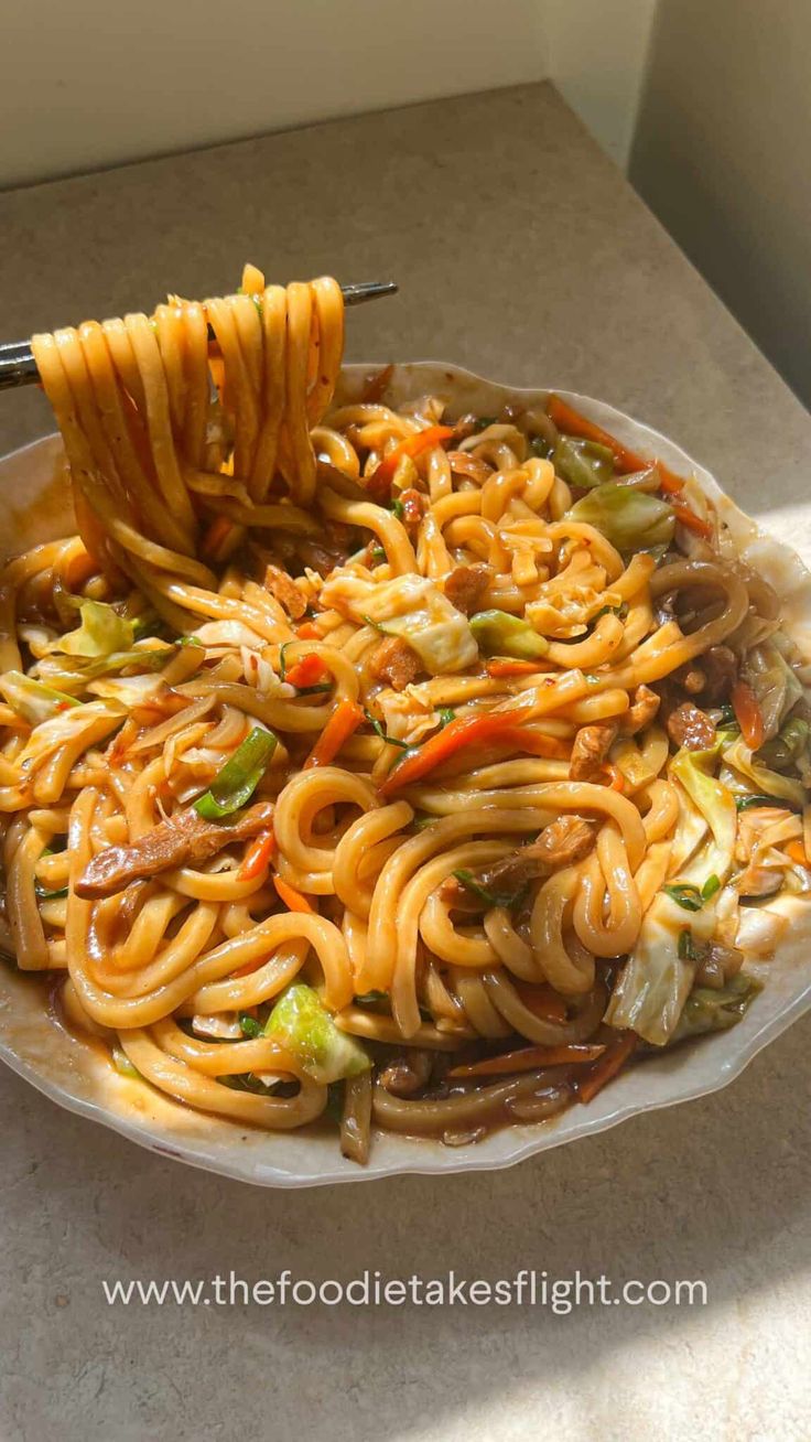 a plate full of noodles and vegetables with tongs sticking out of the top, ready to be eaten