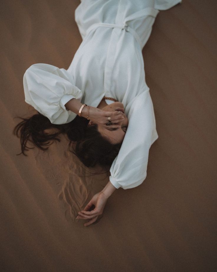 a woman laying in the sand with her hands on her head and wearing a white dress