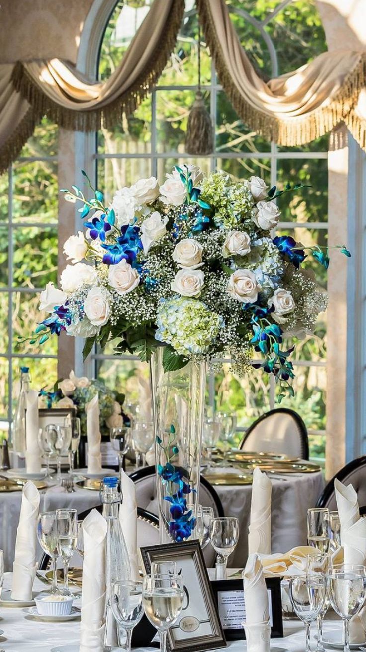 an elegant table setting with blue and white flowers in a tall vase on the centerpiece