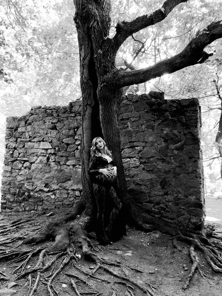 black and white photograph of a woman sitting on a tree stump in front of an old stone building