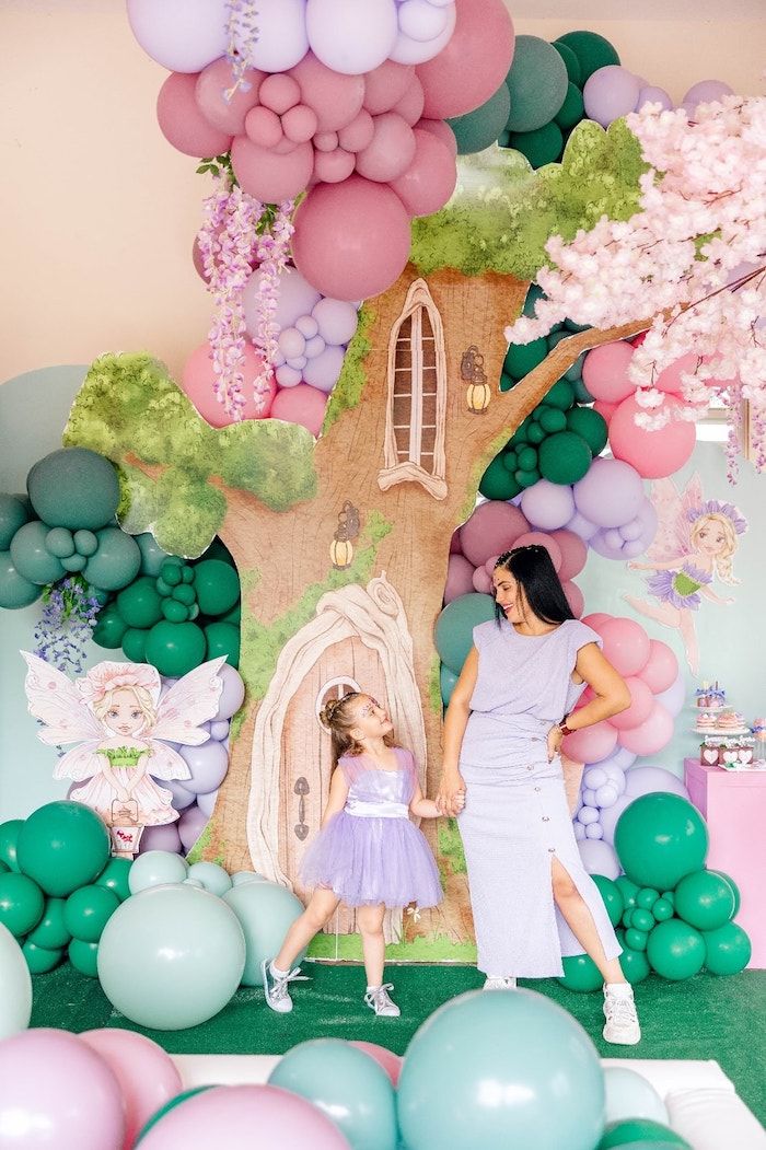 a woman and girl standing in front of a tree with balloons on the wall behind them