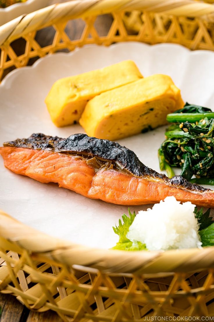 a white plate topped with fish next to green vegetables and rice on top of a wooden table