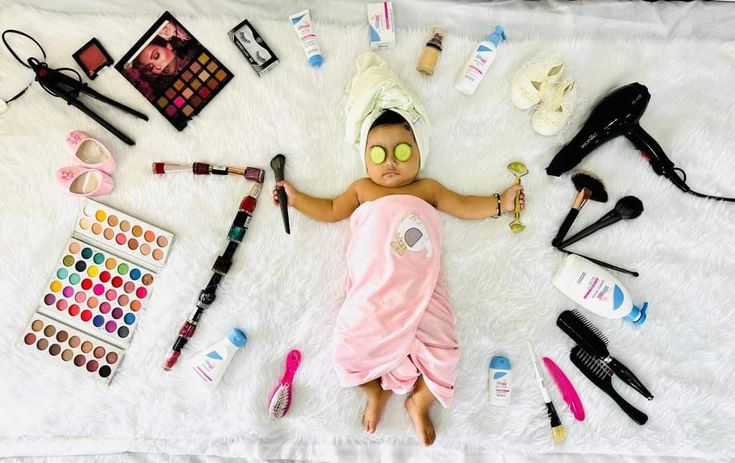 a baby laying on top of a white blanket covered in makeup and other cosmetics items