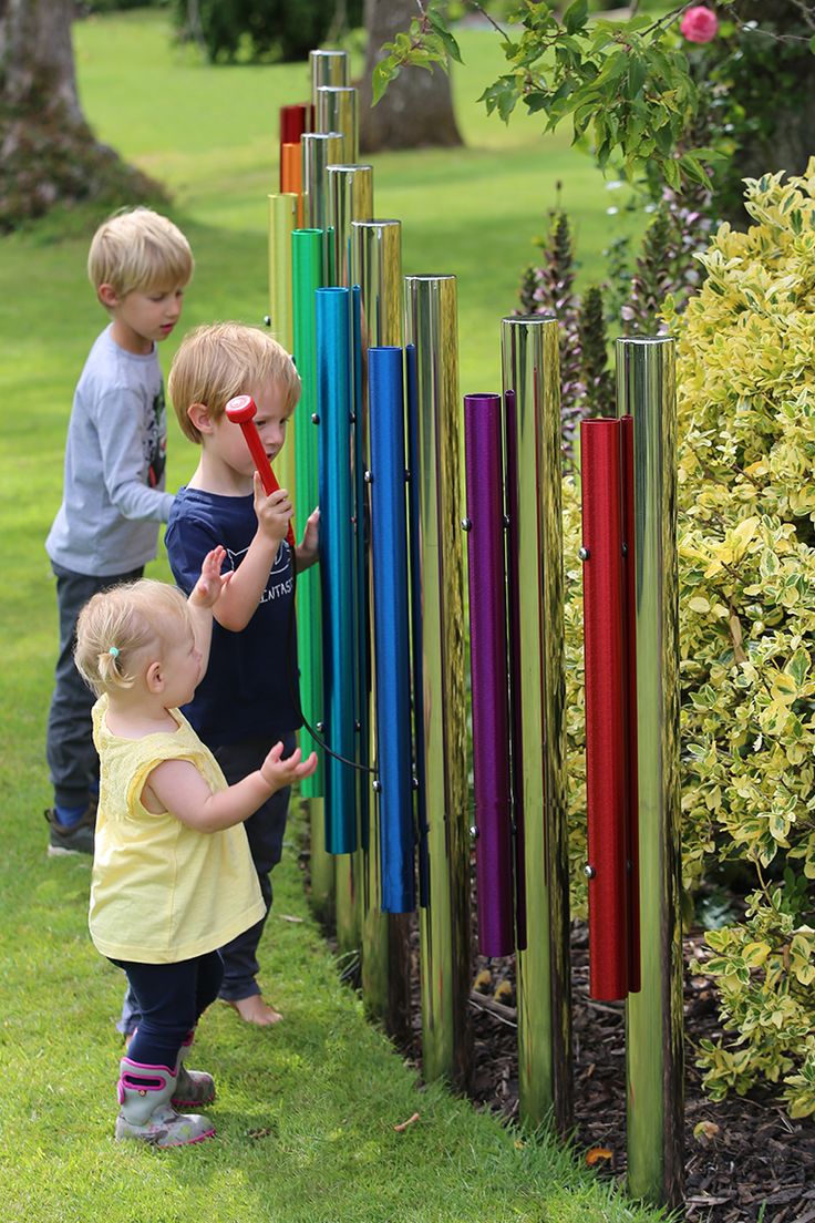 three young children playing with colorful wind chimes in the grass near bushes and trees