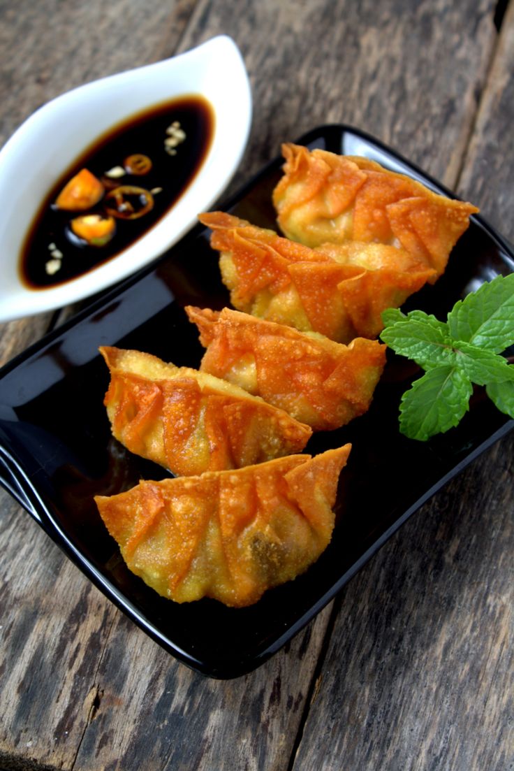 some dumplings are on a black plate next to a bowl of dipping sauce and green leafy garnish