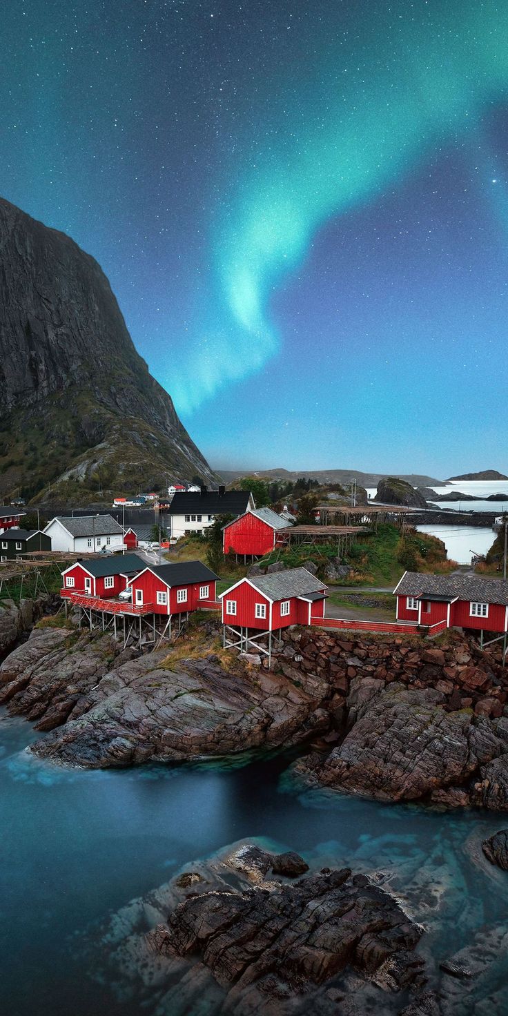 an image of the northern lights in the sky above some red houses and water with rocks