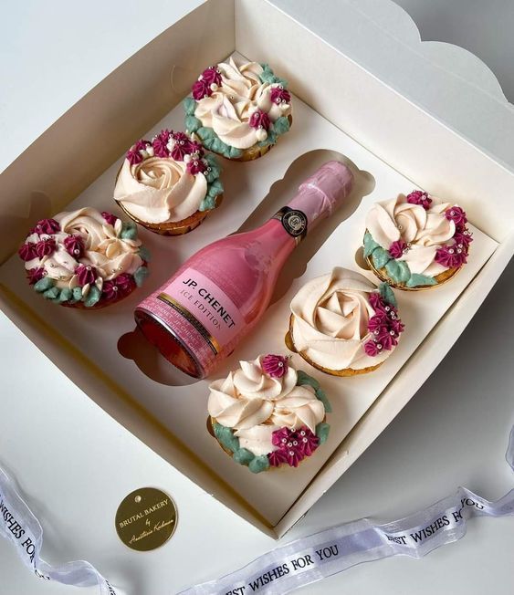 cupcakes with frosting and flowers are in a box next to a bottle