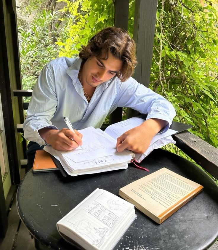 a young man sitting at a table writing in a notebook with an open book beside him