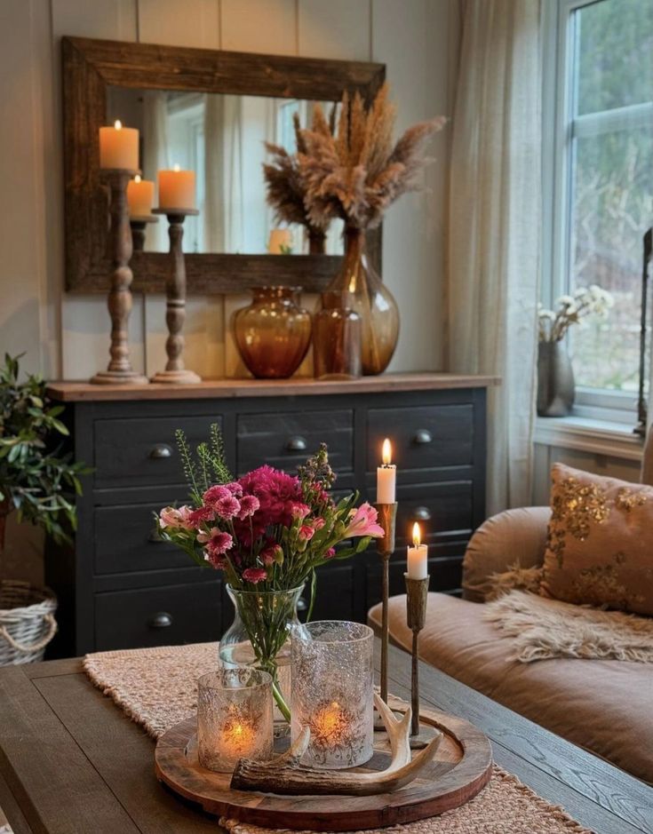 a table with candles and flowers on it in front of a couch, mirror and window