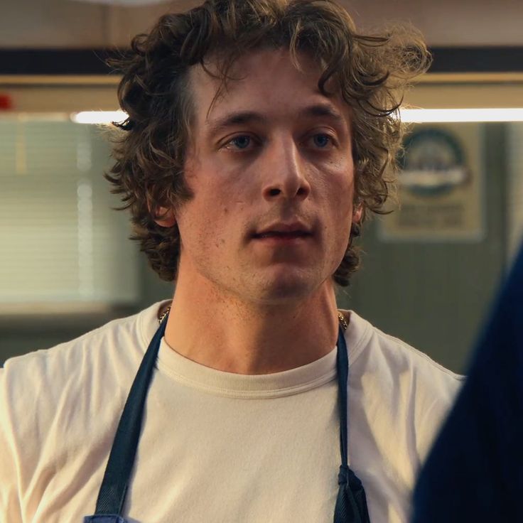 a young man with curly hair wearing an apron and looking at the camera while standing in front of him