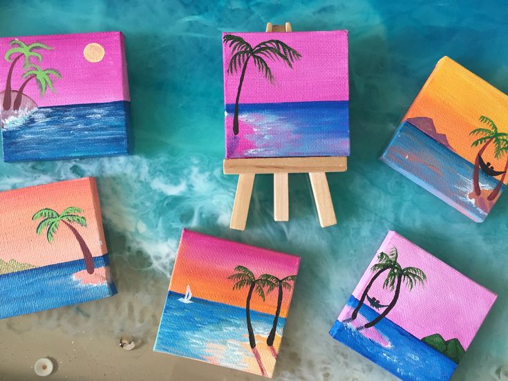 four small canvases with palm trees painted on them sitting in the water next to an easel