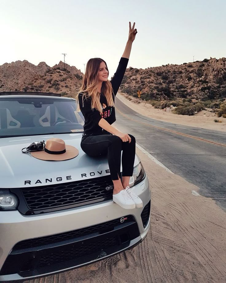 a woman sitting on the hood of a range rover car waving to someone in the desert