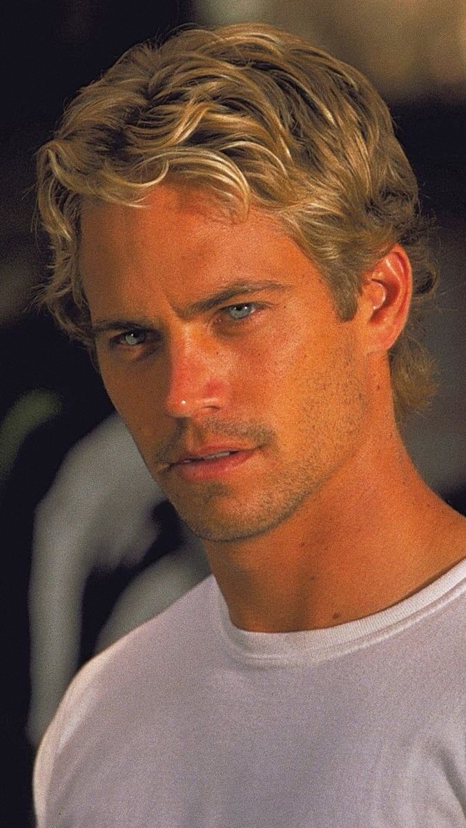 a man with blonde hair and blue eyes looks at the camera while wearing a white t - shirt