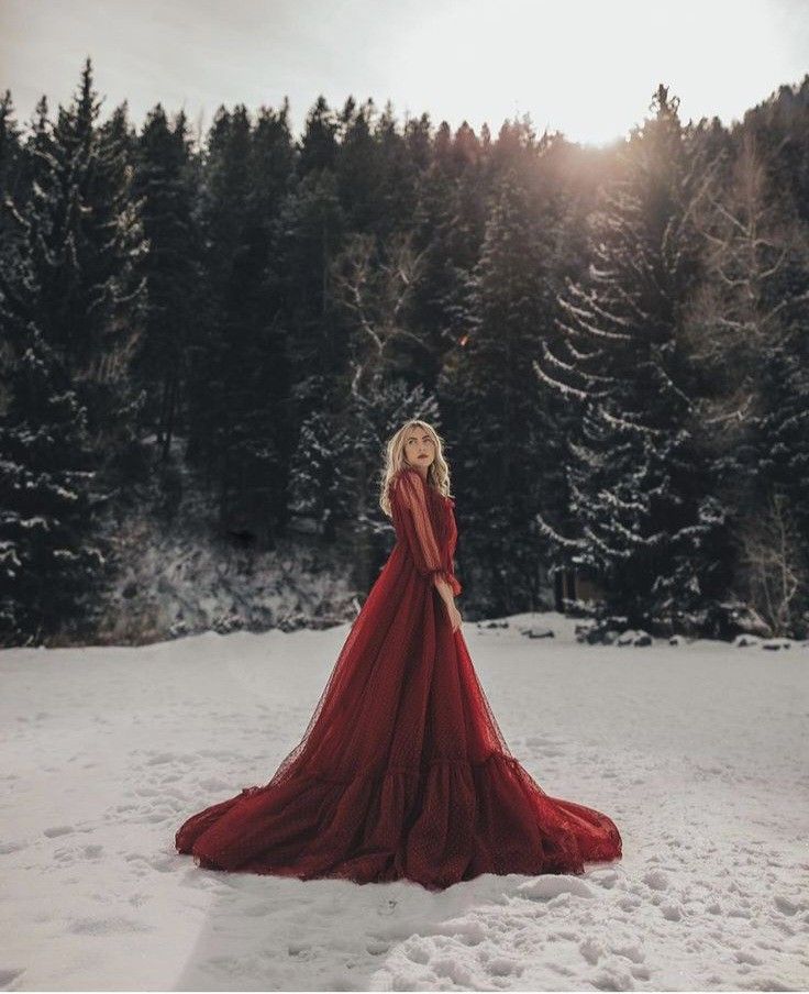 a woman in a long red dress standing in the snow with trees and sun behind her