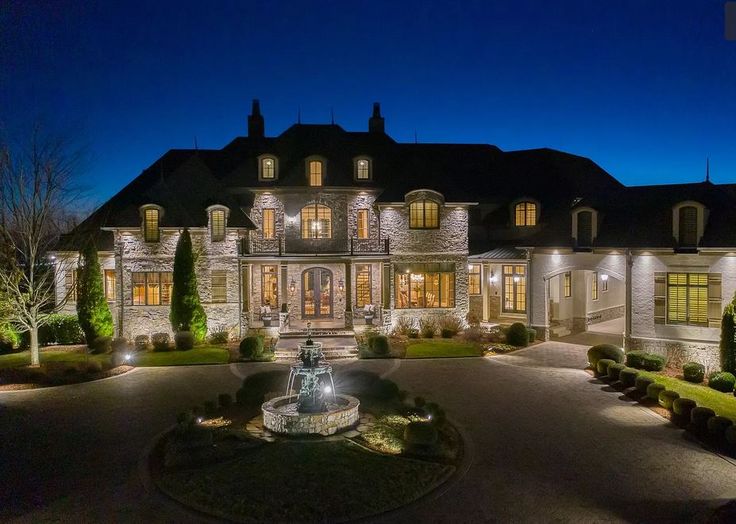 a large house with a fountain in the middle of it at night, surrounded by trees and bushes