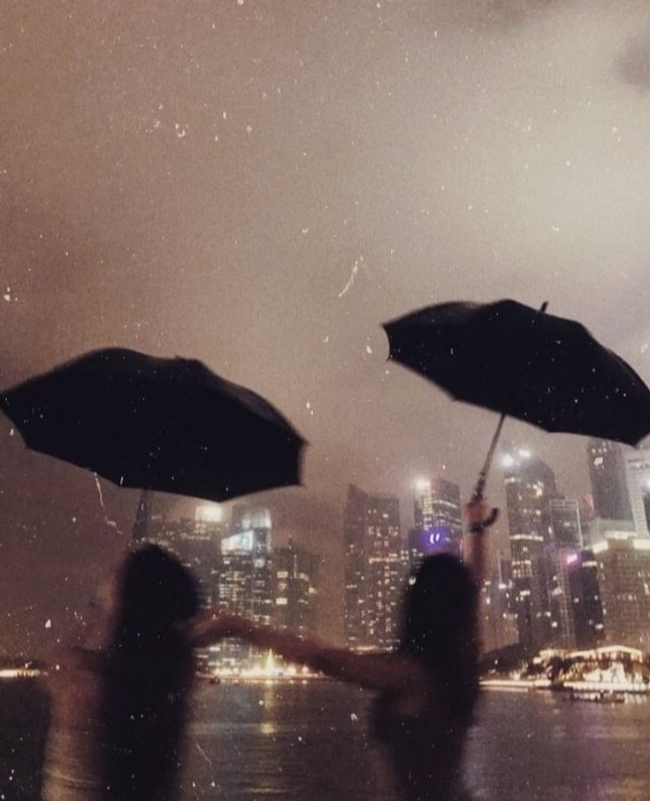 two people holding umbrellas in front of a cityscape at night with rain falling on them