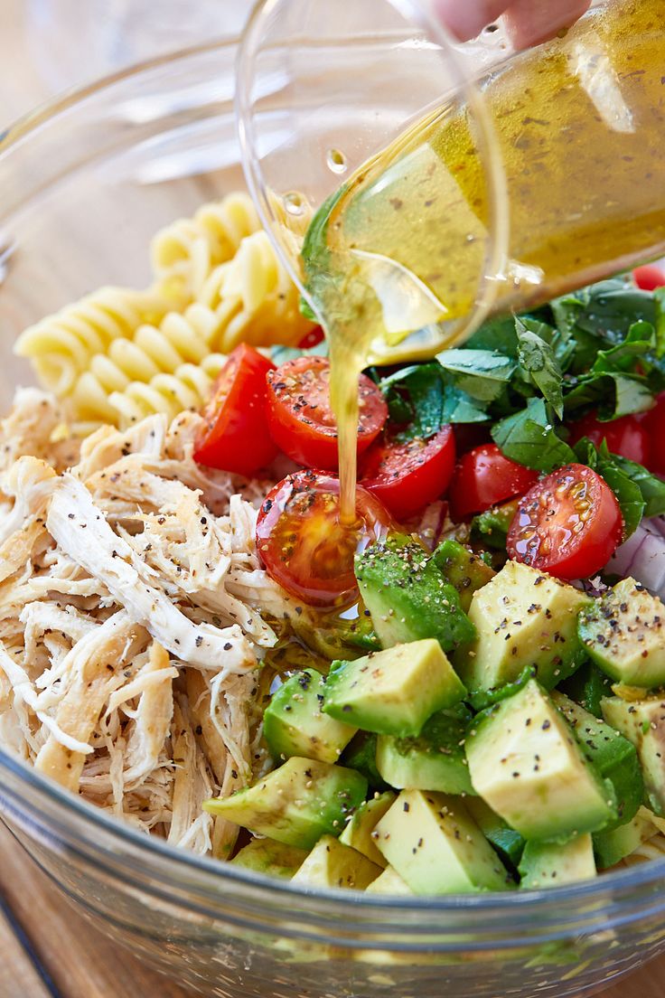 someone pouring dressing into a bowl filled with pasta and veggies, including tomatoes