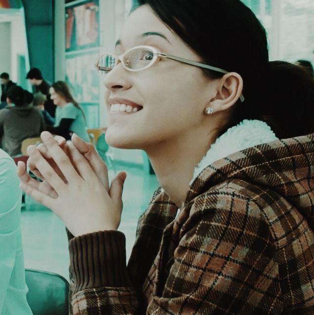 a woman wearing glasses sitting at a table with her hands clasped to her chest and looking off into the distance