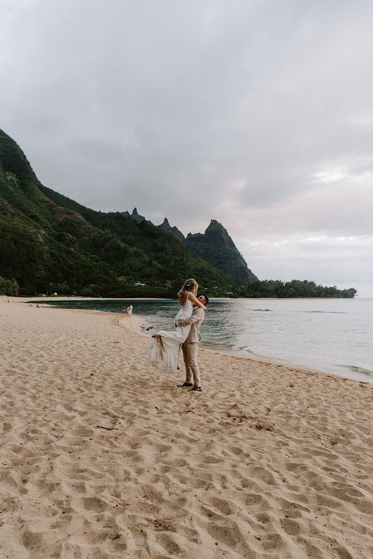 a man and woman standing on top of a sandy beach next to the ocean with mountains in the background