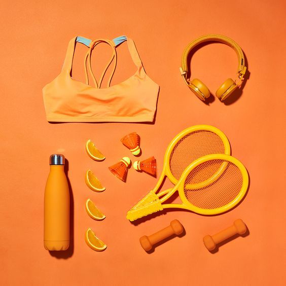 an orange sports bra, headphones, tennis racket, water bottle and other items laid out on a pink surface