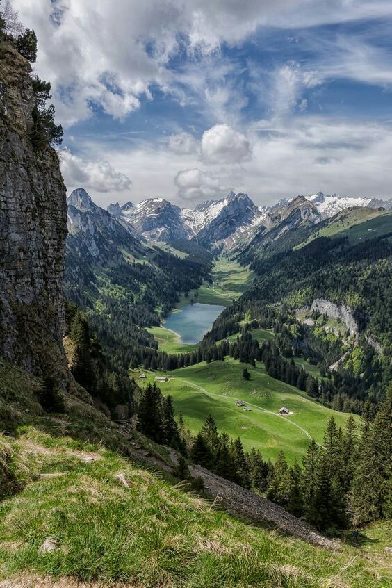 the valley is surrounded by mountains and green grass with trees on both sides, under a partly cloudy sky