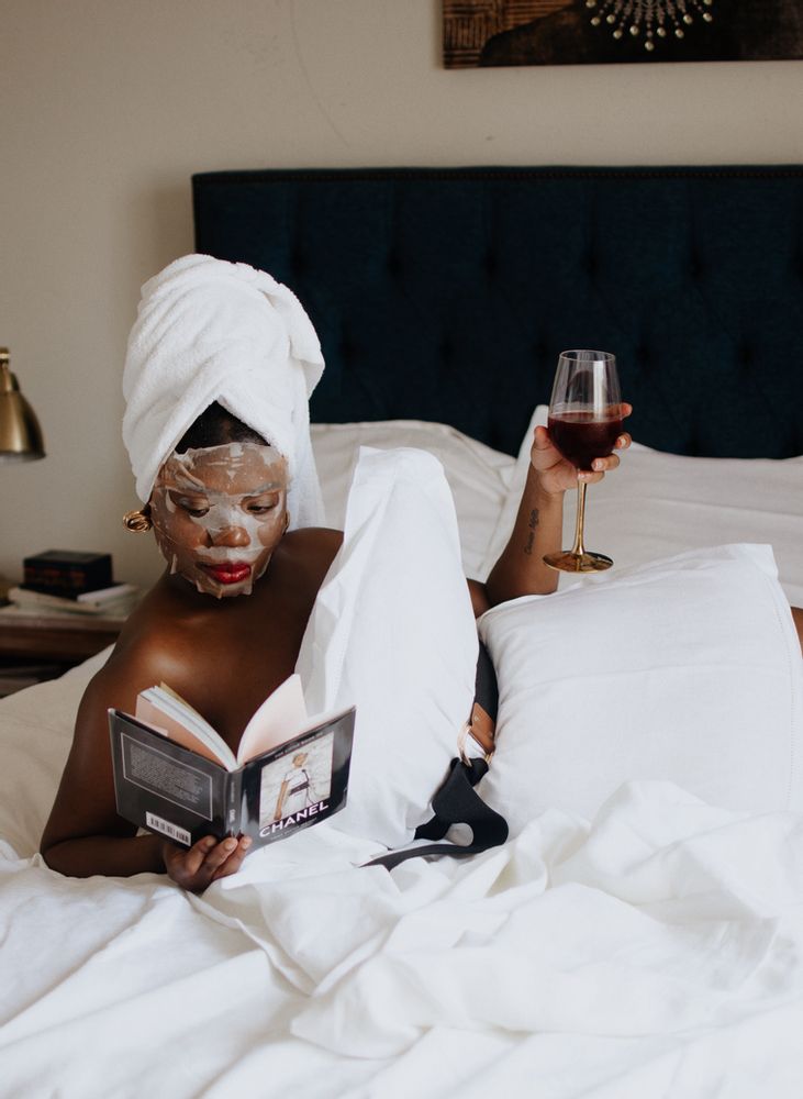 a woman laying in bed reading a book and holding a glass of wine