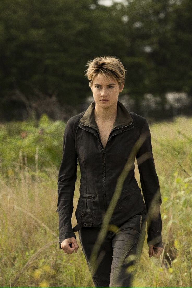 a woman walking through tall grass in a field with her hand on her hip and looking at the camera