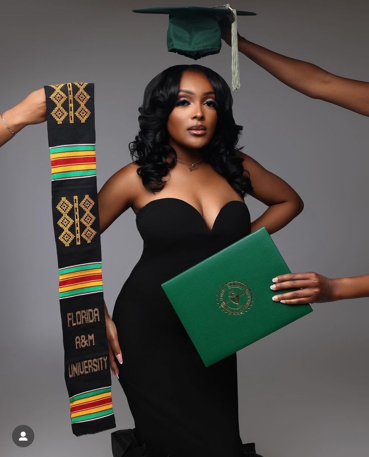 a woman in a black dress holding a green book and wearing a graduation cap with her hands on her head