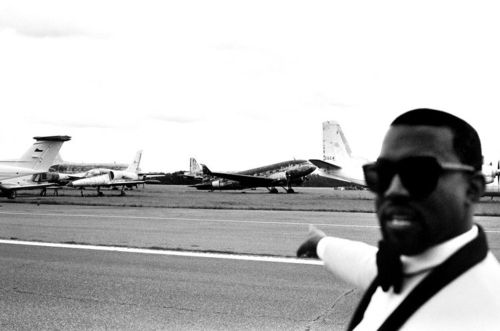 a man in sunglasses and bow tie standing on an airport tarmac with airplanes behind him