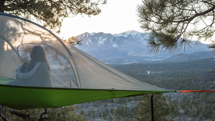 a woman sitting in a hammock on top of a tree covered hill with mountains in the background