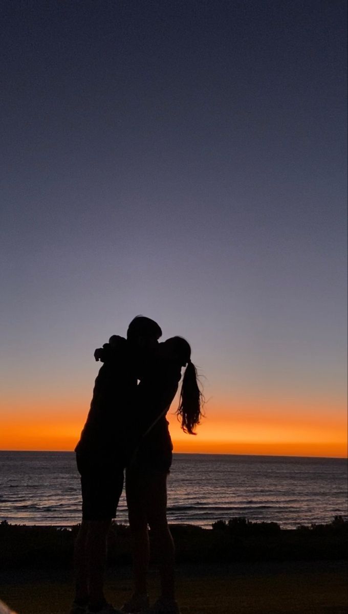 two people standing next to each other in front of the ocean at sunset with their arms around each other