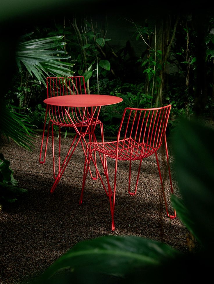 two red chairs sitting next to each other on top of a gravel ground in front of green plants