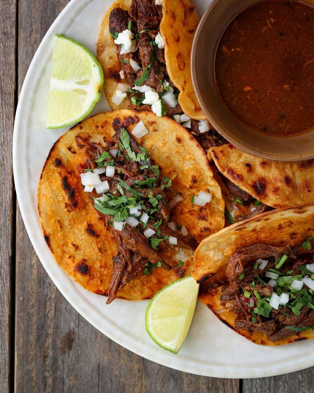 three tacos on a white plate with lime wedges next to it and a bowl of sauce