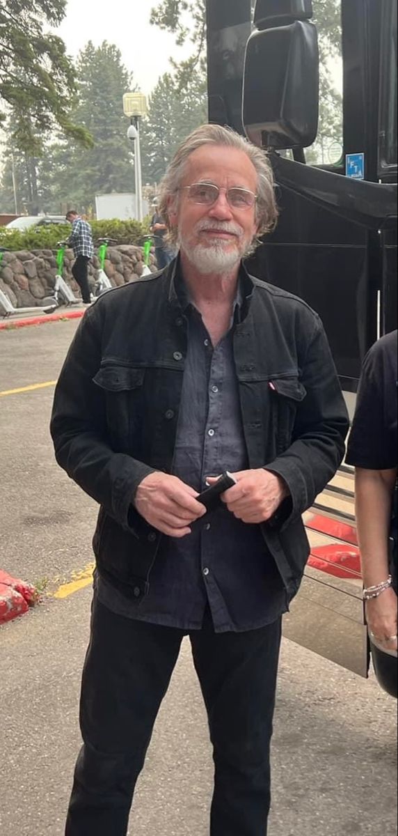 an older man standing in front of a bus with his hands in his pockets and looking at the camera