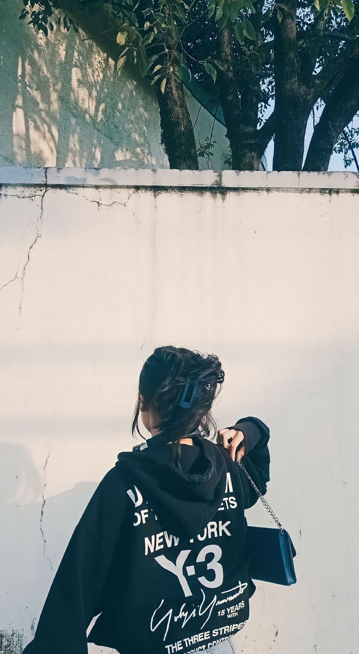 a woman wearing a black hoodie standing in front of a white wall and tree