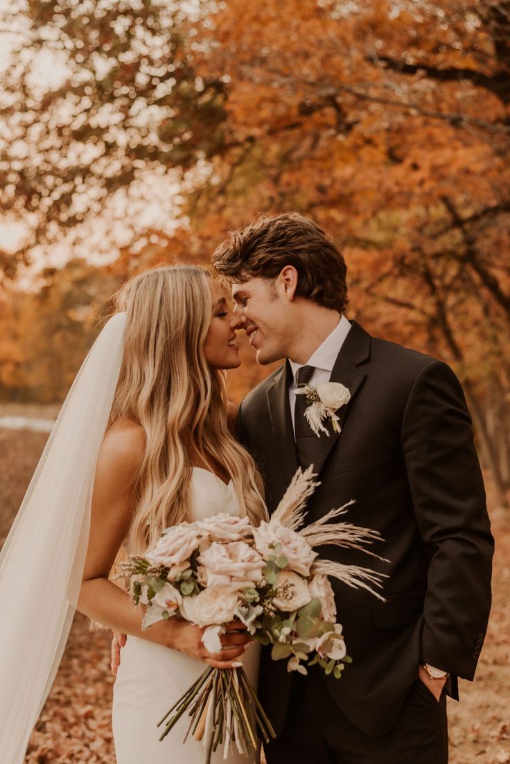 a bride and groom kissing in front of trees with autumn leaves on the ground behind them