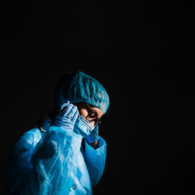 a person wearing a surgical mask and blue coat is standing in the dark with his hands on his face