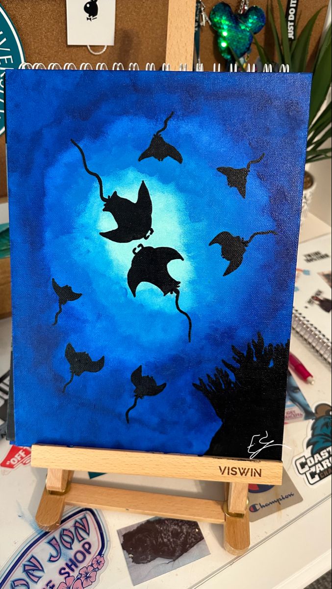 a painting on an easel with blue background and black silhouettes of stingfish